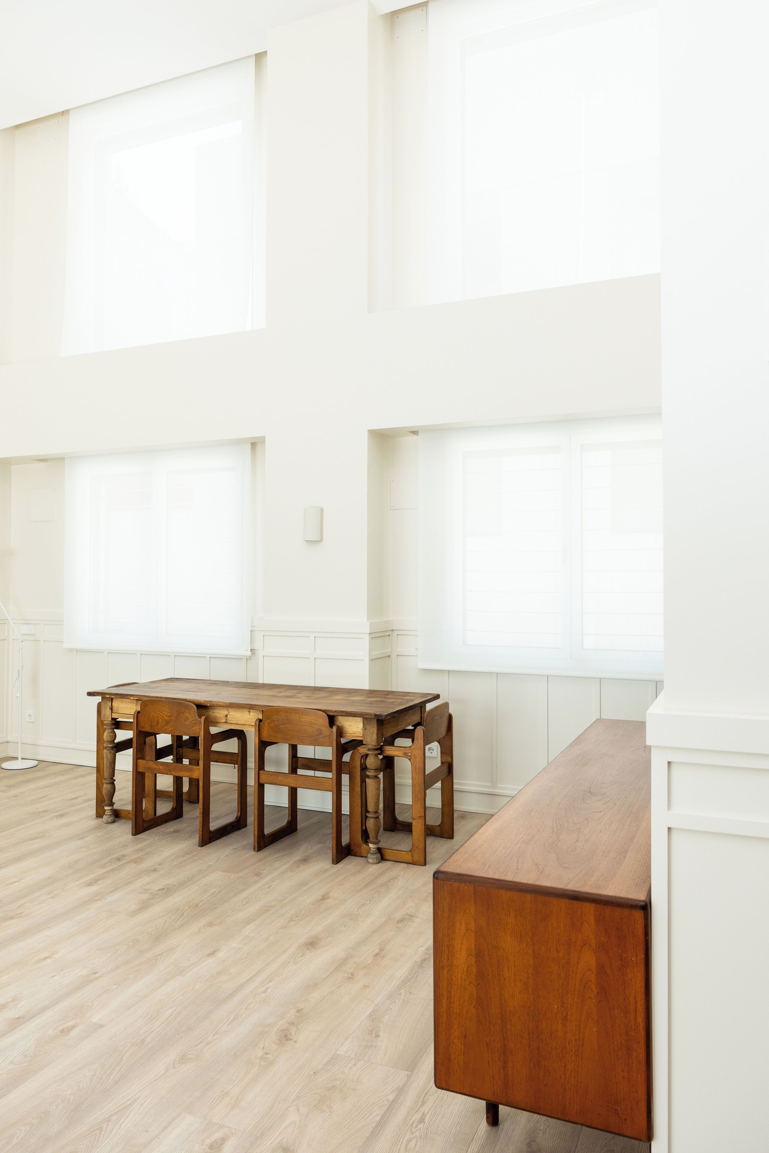 A spacious room with a long wooden table and chairs, large windows, and a white wall.