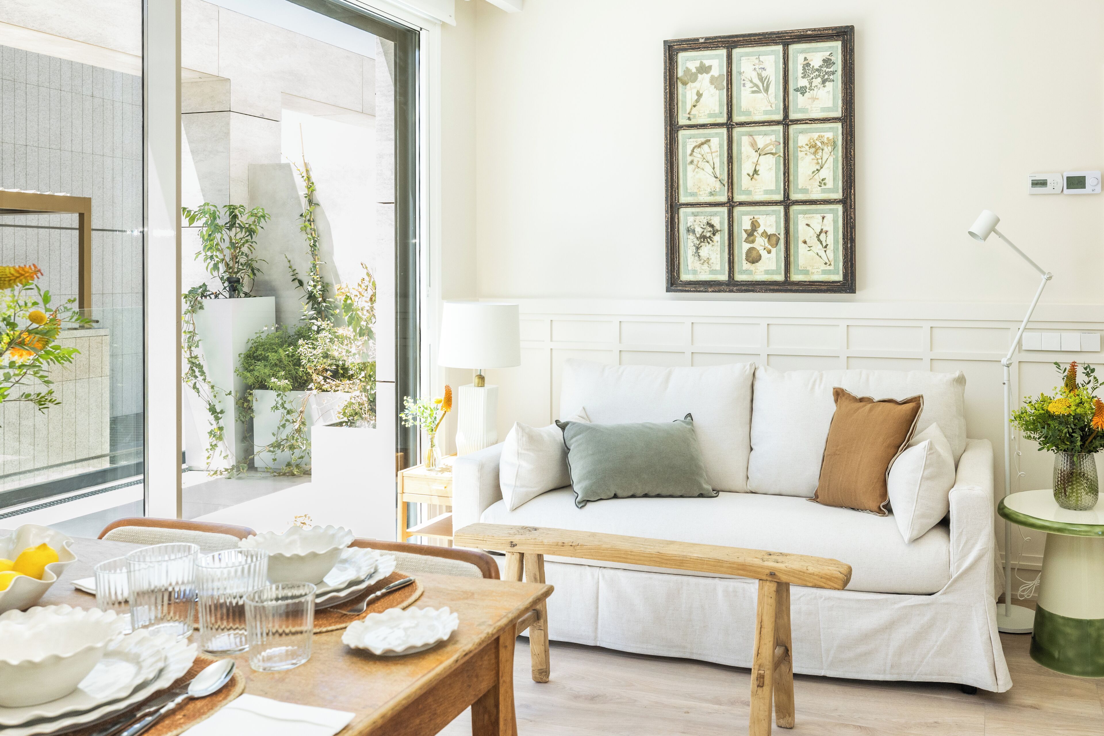 Uma sala de estar moderna com um sofá branco, um banco de madeira e uma mesa de jantar posta com pratos e copos.