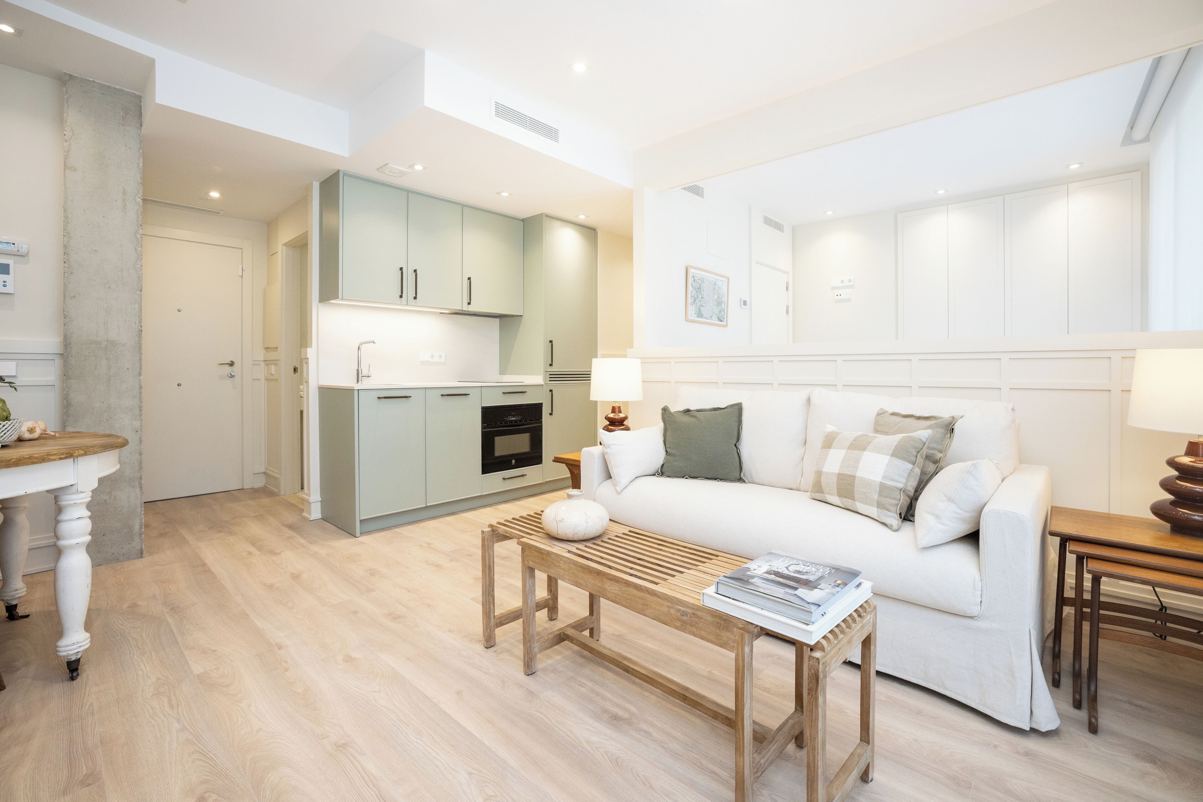 Una sala de estar moderna con un sofá blanco, una mesa de centro de madera y una cocina al fondo.