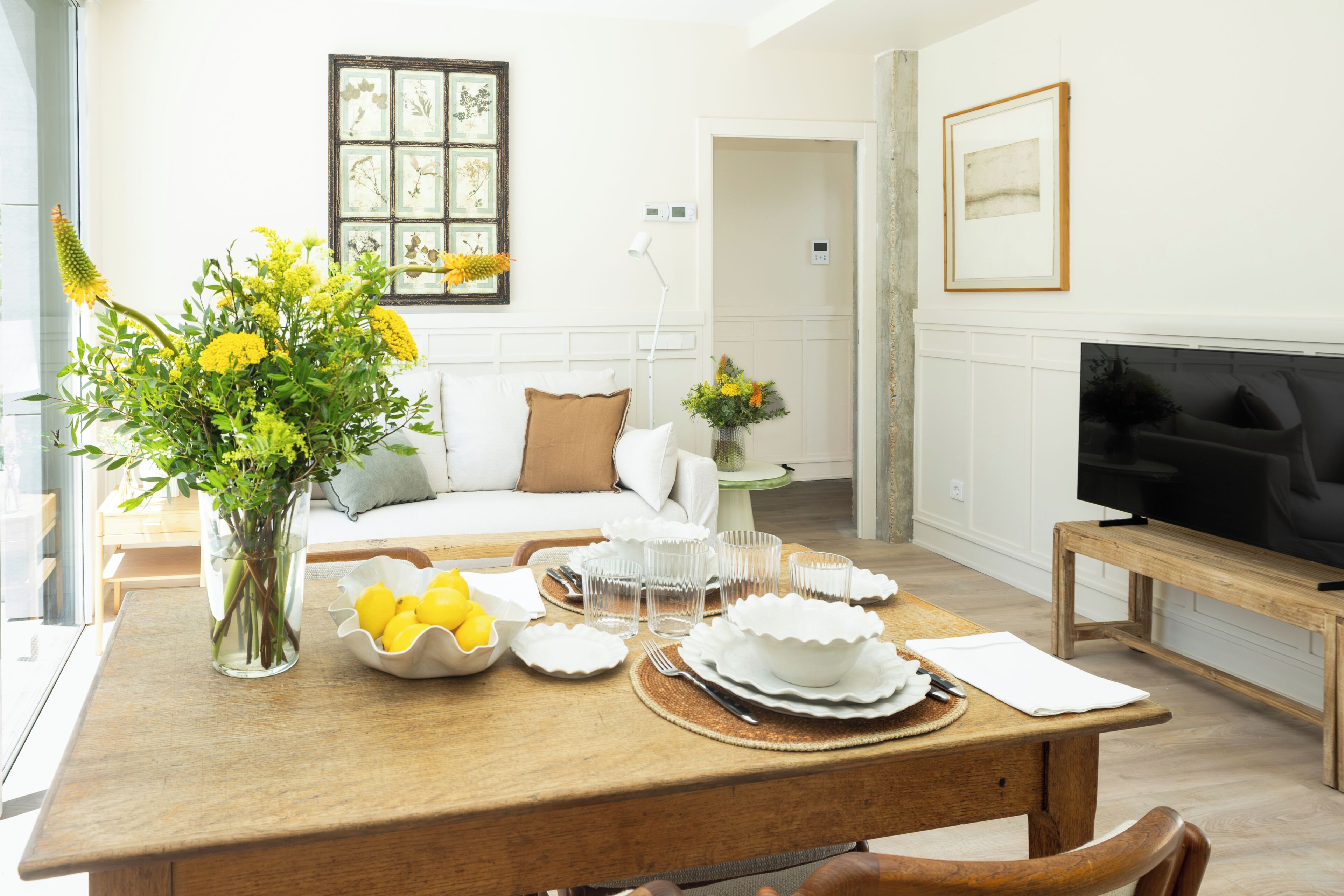 A dining area with a wooden table set for a meal, a vase with yellow flowers, and a television on a wooden stand.
