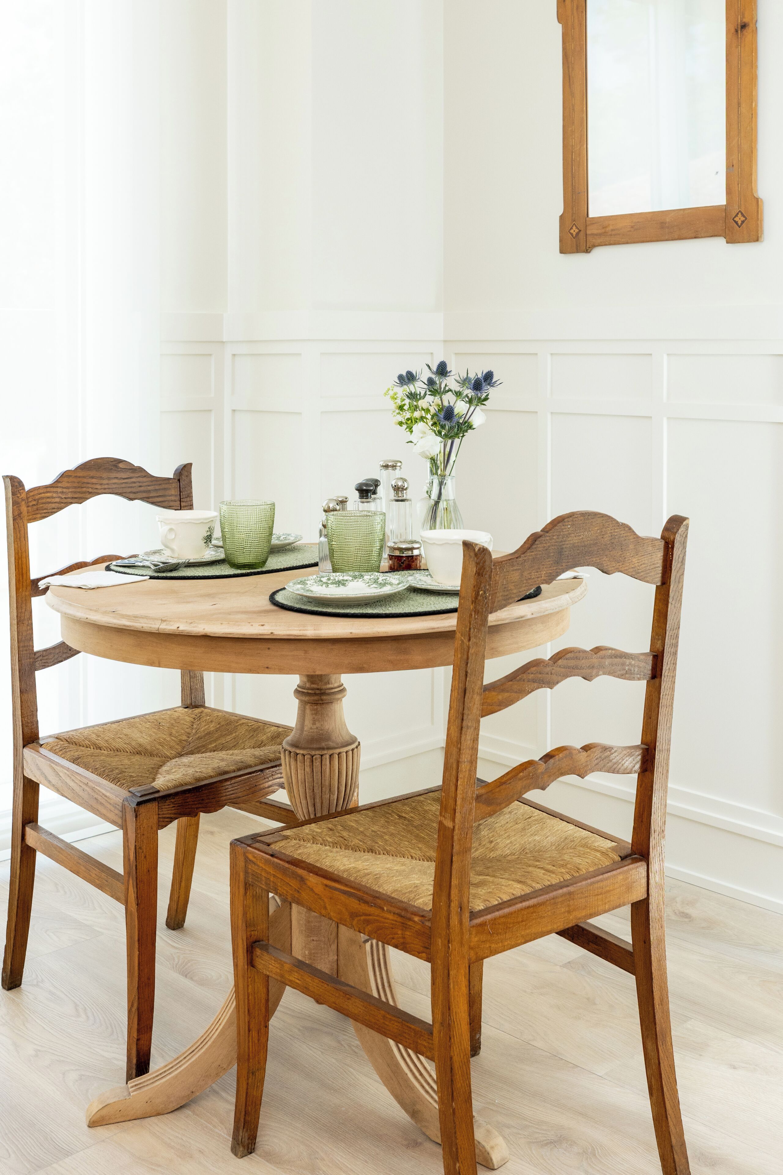 Una mesa de madera redonda con sillas, un florero con flores y diferentes vasos.