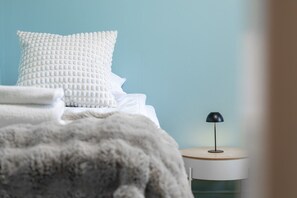 Ein Schlafzimmer mit einem Bett, Kissen, einer Decke, einem Nachttisch mit Lampe und einem zusätzlichen Hocker.