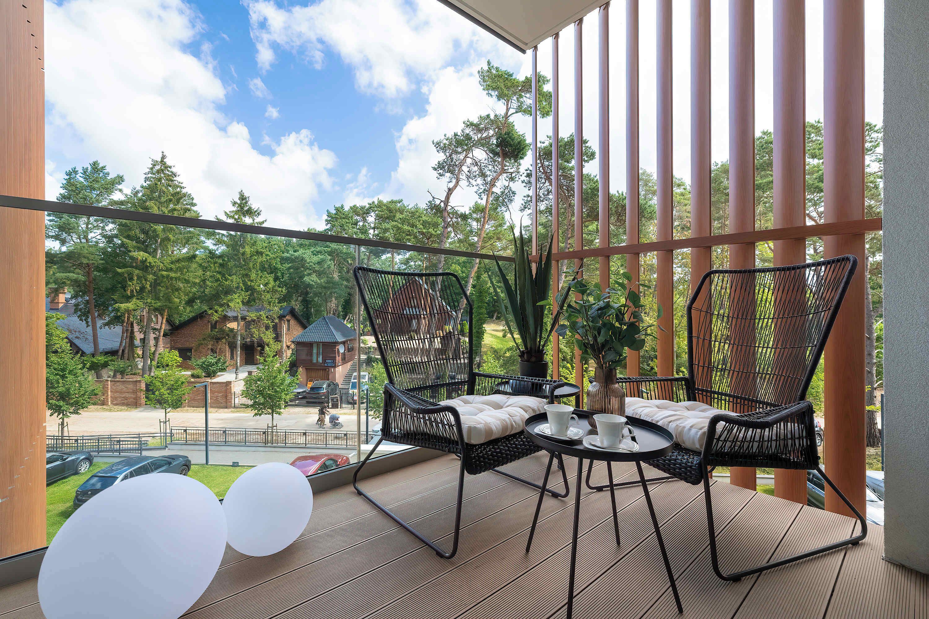 Een balkon met twee rieten stoelen, een klein rond tafeltje en uitzicht op een woonwijk met bomen en huizen.