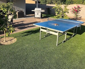 Ein Gartentisch für den Pingpong liegt in einem Hinterhof mit Grill und Gartenmöbeln.