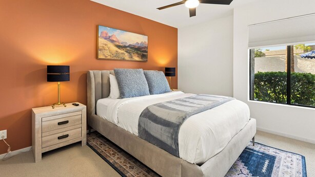 A bedroom with a bed, nightstands, a ceiling fan, a painting, and a window with a view. - Elevated Desert Escape | Heated Pool • Putt Green (Scottsdale)
