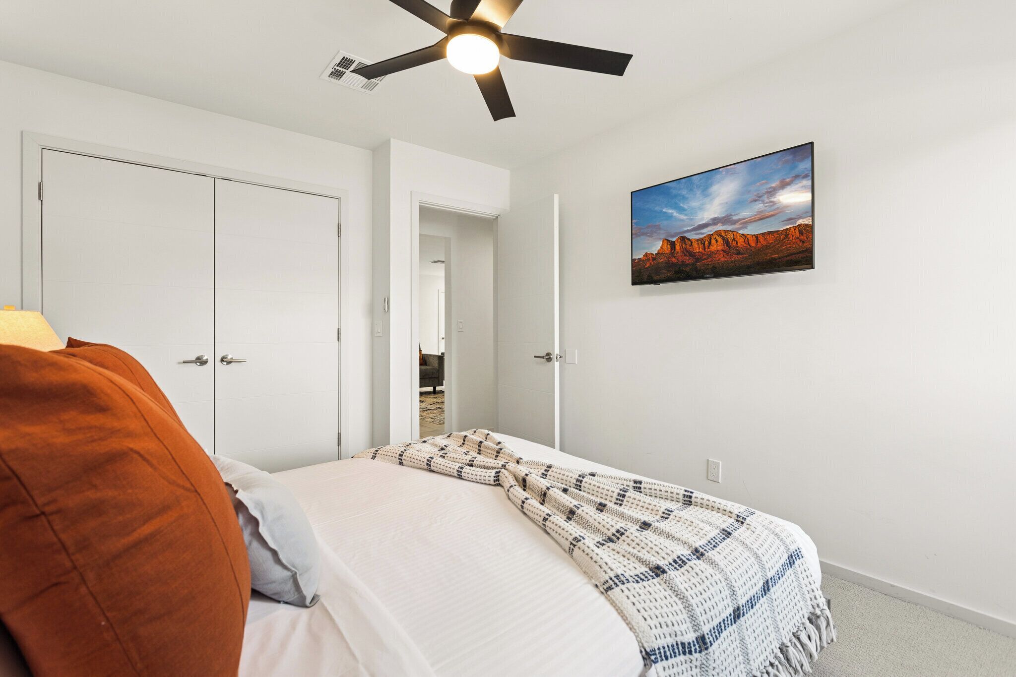 A bedroom with a bed, a ceiling fan, a wall-mounted TV, and a door leading to another room.