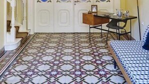 Una habitación con piso estampado, una puerta blanca con paneles de vidrio, un candelabro, una mesita con una lámpara y una cama con cubrecama estampado.