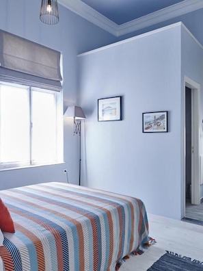 Ein Schlafzimmer mit einem Bett, einem Fenster mit Vorhängen, einer Lampe und zwei gerahmten Bildern an der Wand.