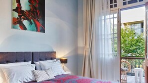 Quarto com cama, mesa de cabeceira, luminária e uma janela grande com cortinas.