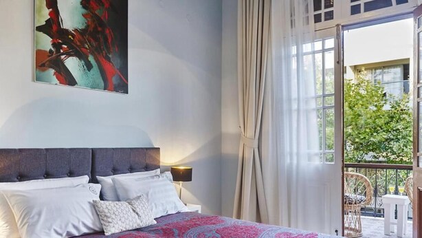 Quarto com cama, almofadas, mesa de cabeceira, luminária e uma janela grande com cortinas.