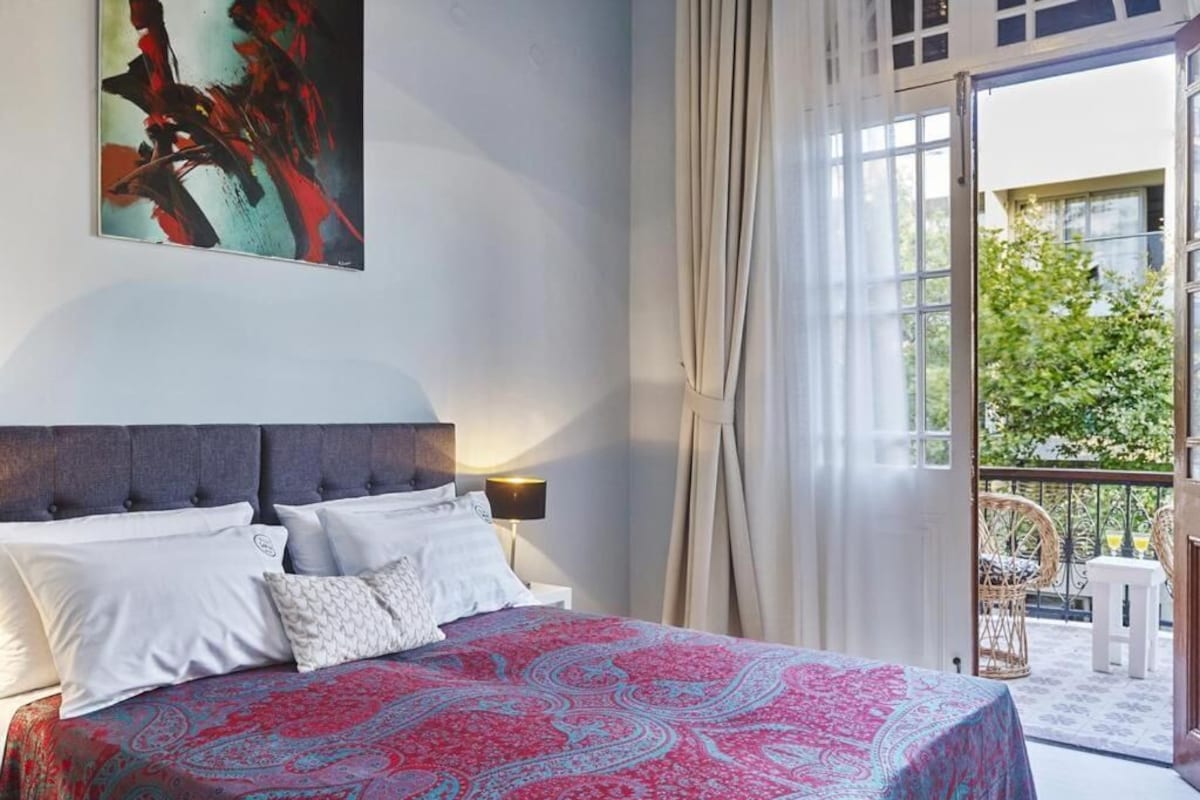 Ein Schlafzimmer mit einem Bett, Kissen, einem Nachttisch, einer Lampe und einem großen Fenster mit Vorhängen.