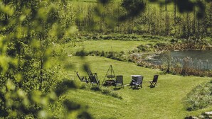 Un espacio al aire libre con césped, un estanque rodeado de árboles y vegetación. Hay varias sillas y una mesa dispuestas para comer al aire libre.