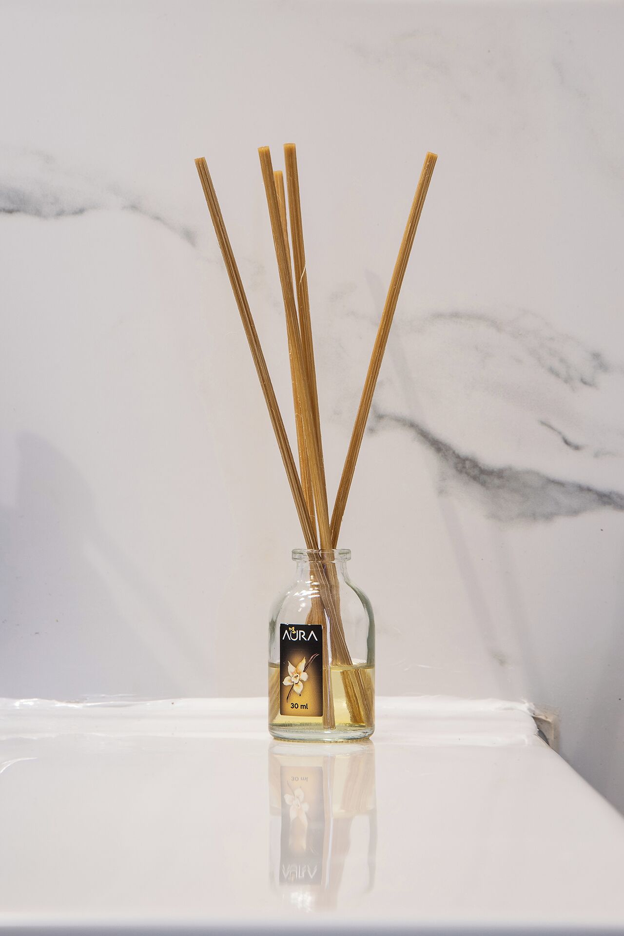 A glass bottle with reed sticks and a label, placed on a reflective surface.
