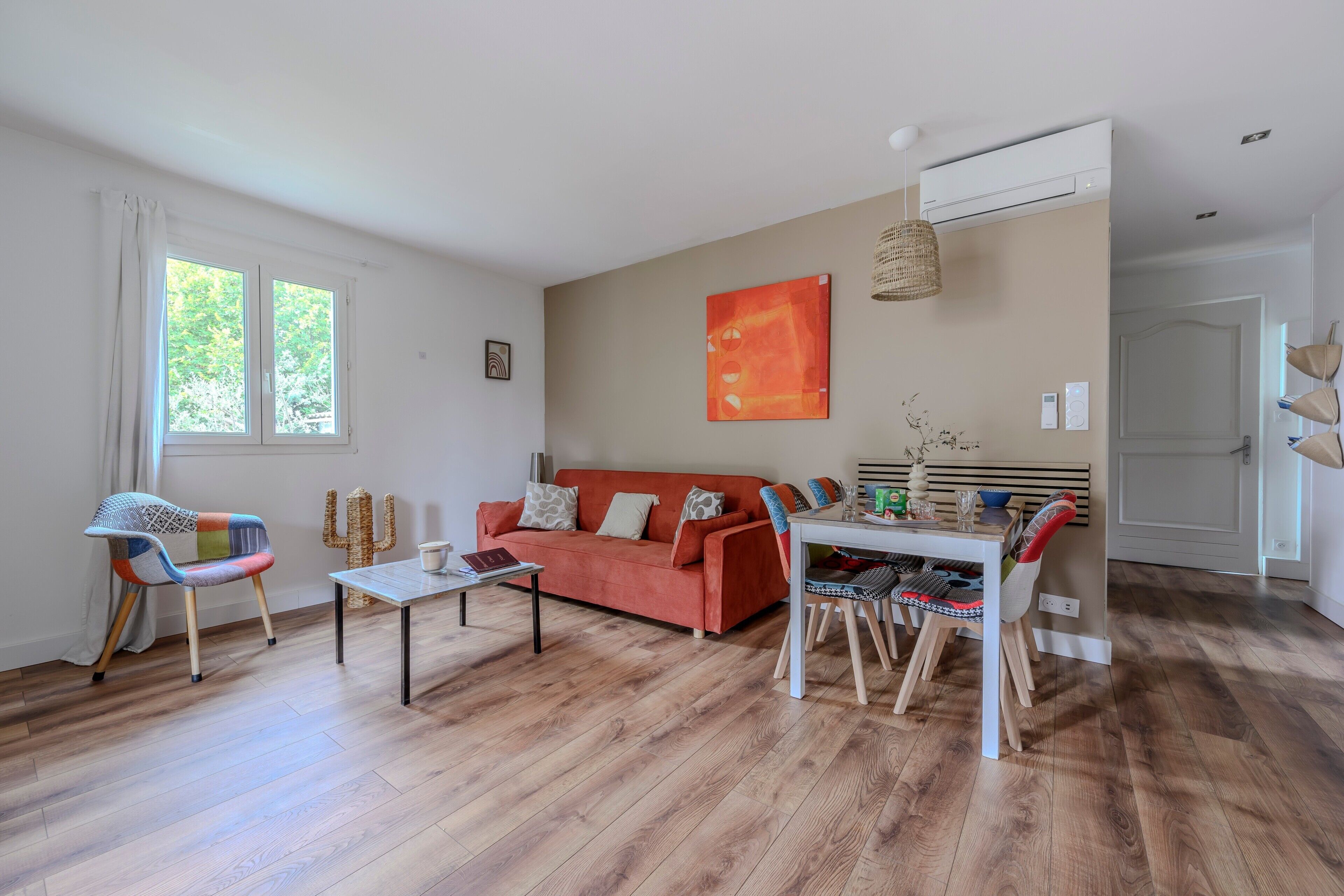 Sala de estar con un sofá, una mesa de centro y una mesa de comedor con sillas. Hay una ventana con cortinas, un aire acondicionado de pared y un cuadro.