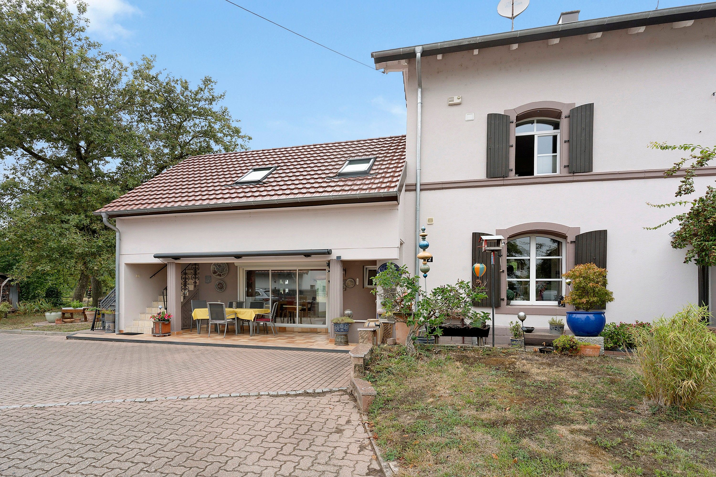 Ein zweistöckiges Haus mit Ziegeldach, einer überdachten Terrasse mit Gartenmöbeln und einem Garten mit Blumentöpfen.