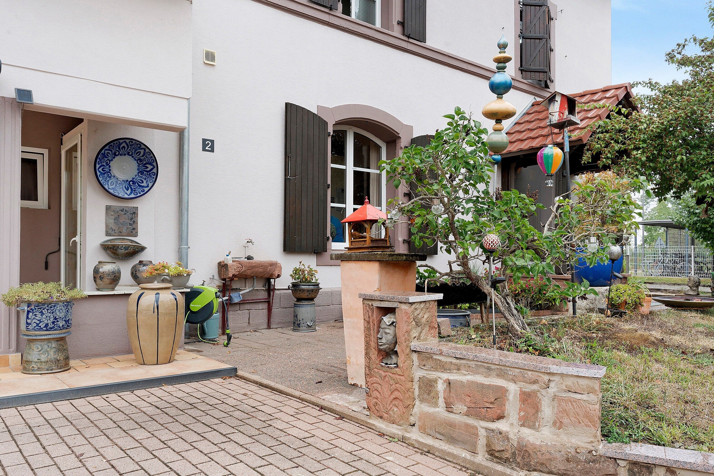 Un patio con suelo de baldosas, plantas en macetas, un muro de piedra y un edificio con puerta y ventana.