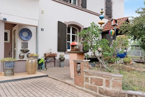 A courtyard with a tiled floor, potted plants, a stone wall, and a building with a door and window.
