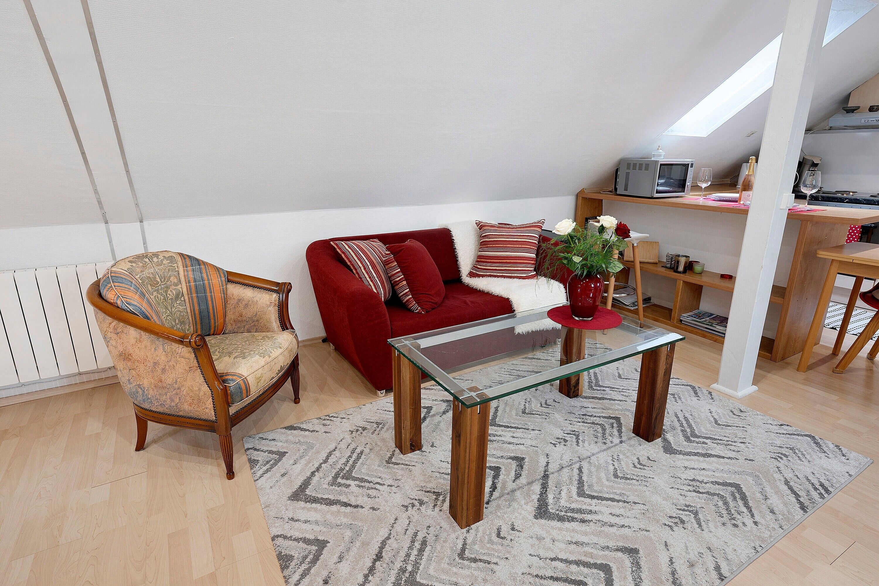 Una sala de estar acogedora con un sofá rojo, un sillón de madera, una mesita de centro de vidrio y un comedor con un microondas y una botella de vino.