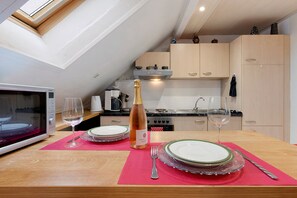 A kitchen with a dining table set for two, a microwave, and a bottle of champagne.
