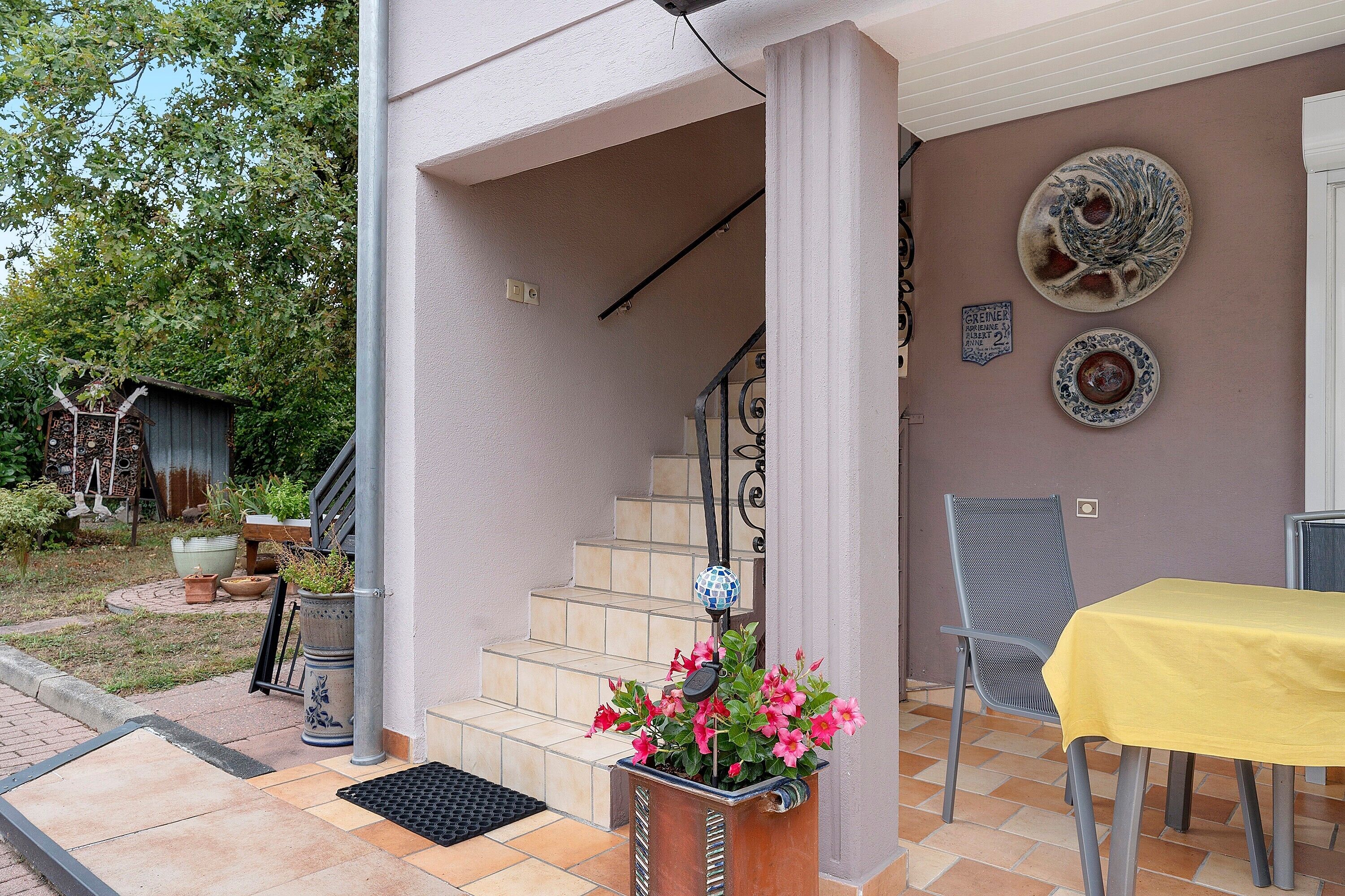 Ein überdachter Außenbereich mit einer Treppe, die zu einer zweiten Etage führt, einem Metallstuhl, einem gelben Tisch und einem Blumentopf mit rosa Blüten.