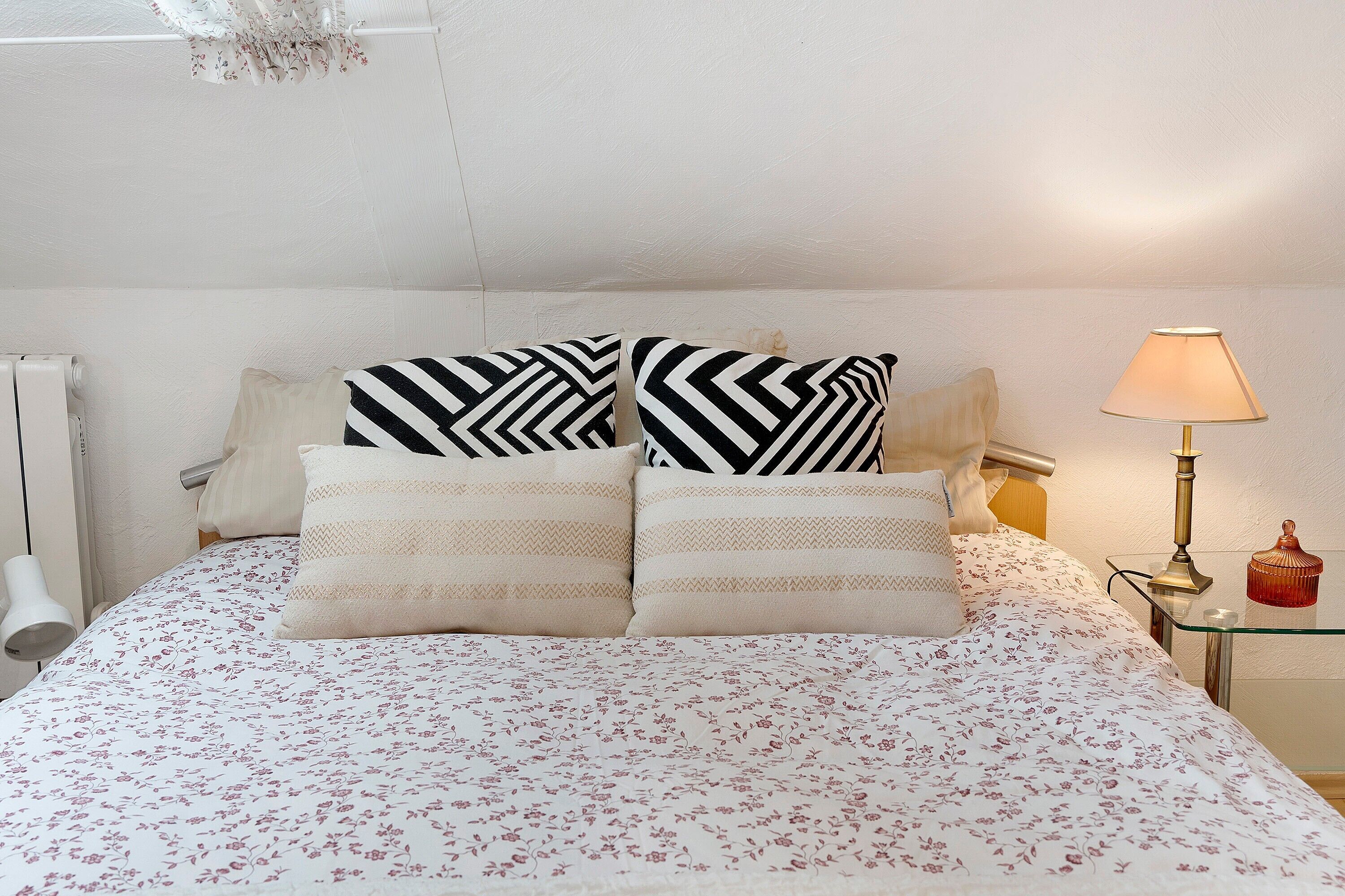 Un lit avec une literie à motifs floraux, deux coussins noirs et blancs à rayures, une lampe de chevet et un petit objet rouge posé sur la table de chevet.