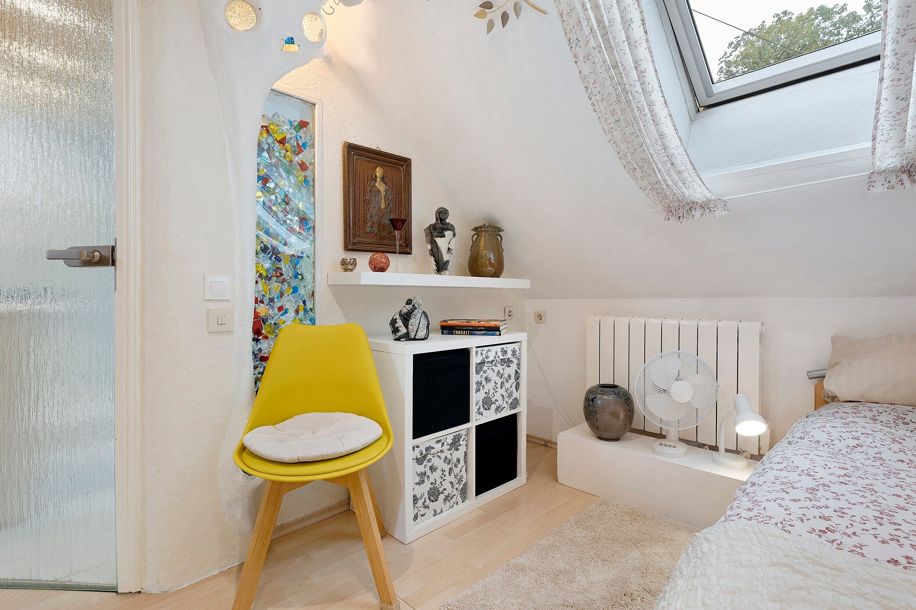 Ein Schlafzimmer mit einem Bett, einem gelben Sessel, einer weißen Kommode und einem Fenster mit Blick auf Bäume.
