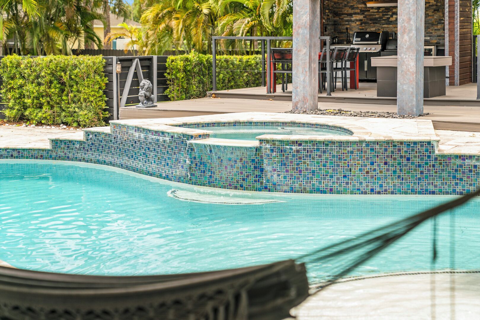 A swimming pool with a mosaic tile border, surrounded by a landscaped area with palm trees and a barbecue setup.
