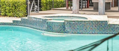A swimming pool with a mosaic tile border, surrounded by a landscaped area with palm trees and a barbecue setup.