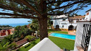 Zona de piscina con vista al mar, rodeada de vegetación y un edificio blanco con tejado de tejas.