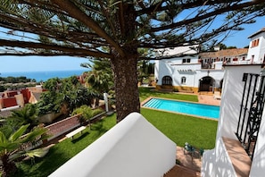 Ein Poolbereich mit Blick auf das Meer, umgeben von Grünflächen und einem weißen Gebäude mit einem Ziegeldach.