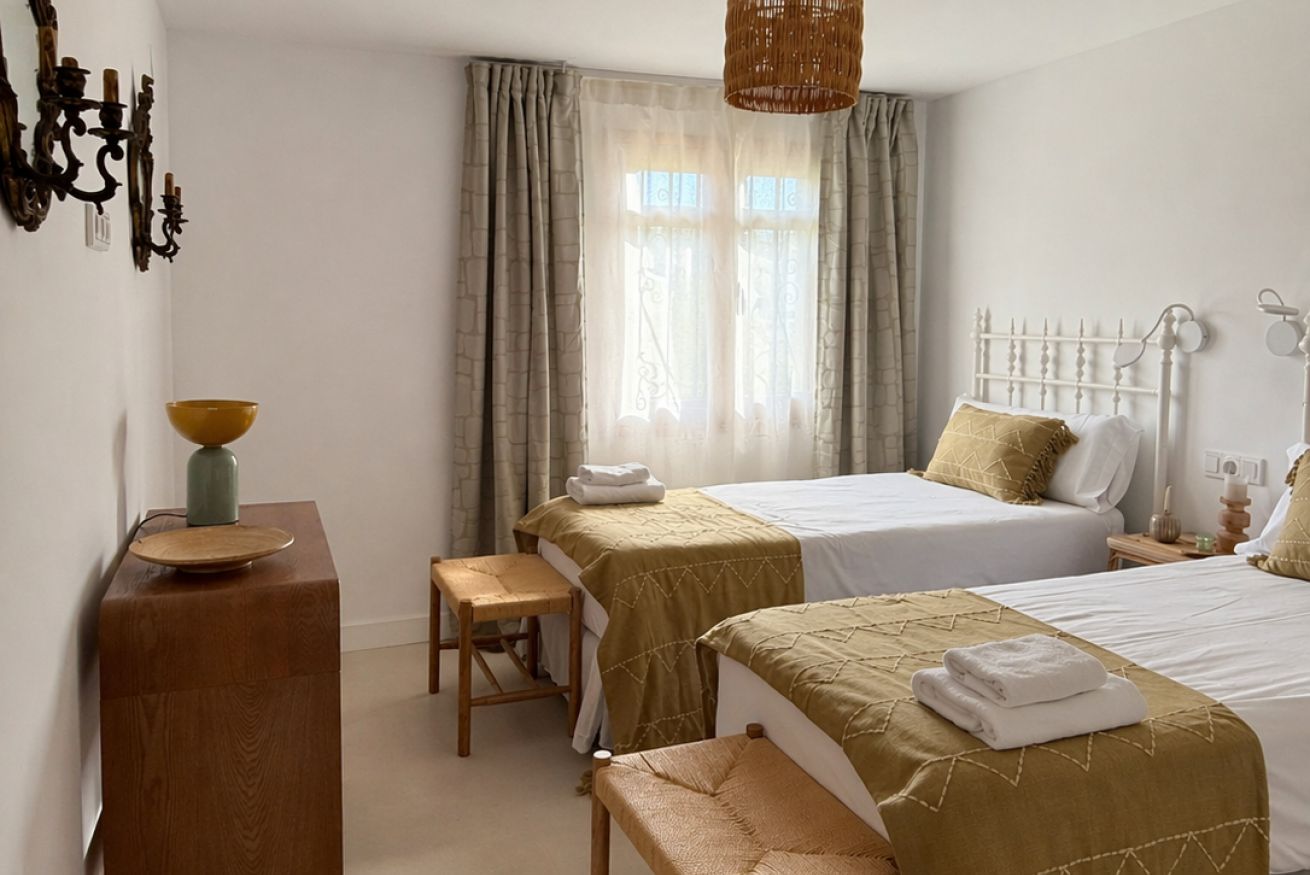 Quarto de hotel com duas camas, uma escrivaninha de madeira, uma cadeira, uma mesinha de cabeceira e uma janela com cortinas.