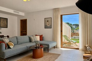 Ein modernes Wohnzimmer mit einem Ecksofa, einem Couchtisch und Blick auf eine grüne Rasenfläche sowie Gebäude durch eine Glastür.