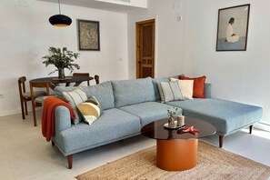 Ein Wohnzimmer mit Ecksofa, rundem Couchtisch, Esstisch mit Stühlen und Bildern an den Wänden.