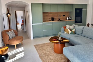 A modern living room with a sofa, armchair, coffee table, and a kitchen area with cabinets and appliances. - Ten Pool Apartment at Hacienda Clavero (Málaga)