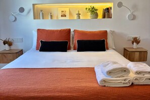 Una cama bien tendida con dos almohadas, una mesita de noche con un florero, y un estante de pared con adornos.