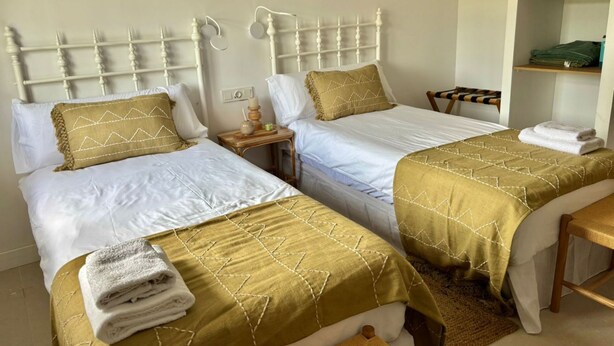 Dos camas individuales con cabeceros blancos y ropa de cama dorada, una mesita de noche con lámpara y un banco de madera.