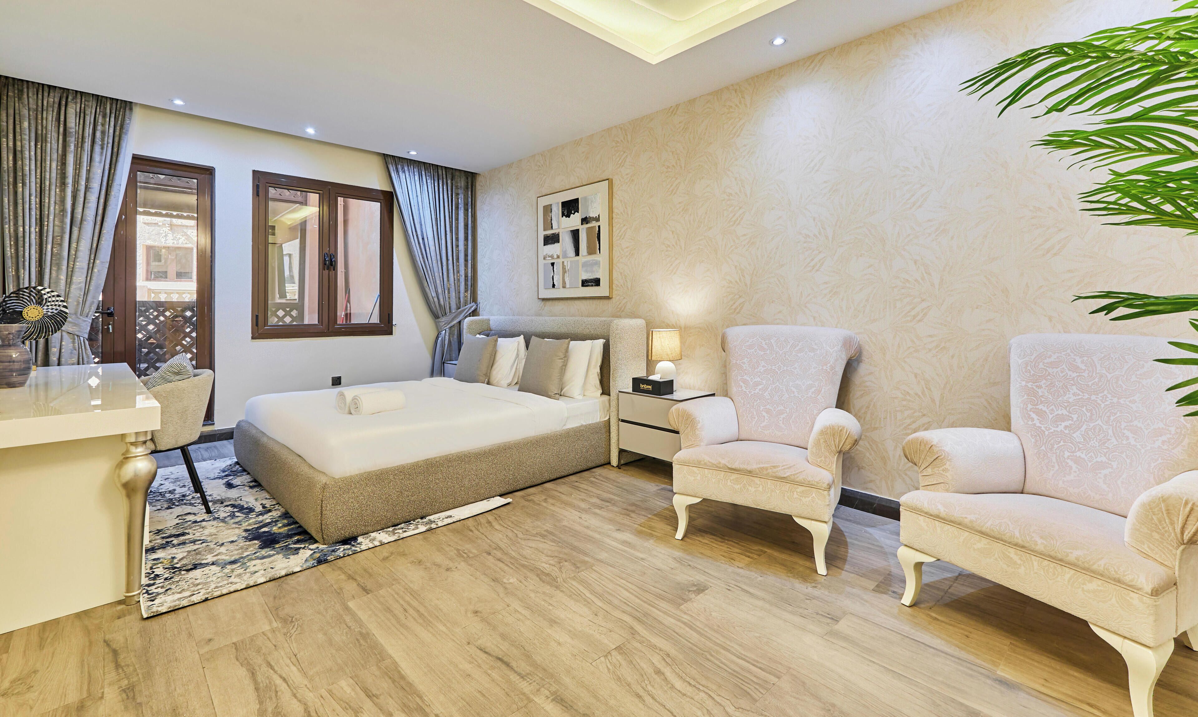 Une chambre d’hôtel avec un lit, deux fauteuils, un bureau et une grande fenêtre avec des rideaux.