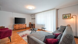 A modern living room with a grey sofa, red and black cushions, a flat-screen TV, wooden cabinet, and a large window with sheer curtains. - Holiday Home Cerine Hill by Villas Guide (Gornji Vinkovec)