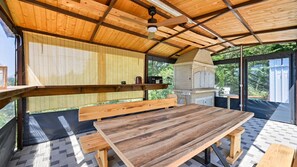 Eine Terrasse im Freien mit Holztisch und -bänken, einem Backofen aus Ziegeln und einem Deckenventilator.