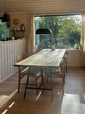 A dining area with a wooden table and chairs, a hanging pendant light, and a view of greenery through a window.