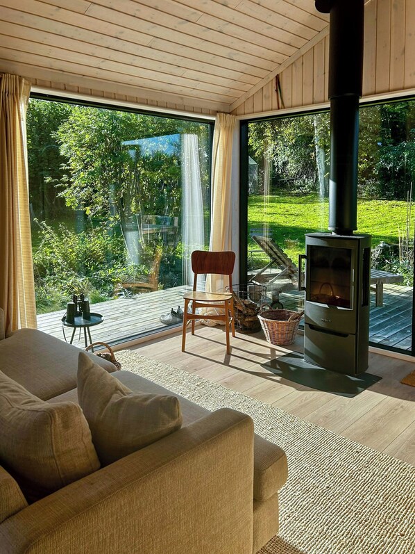 A modern living room with a sofa, a wooden chair, a fireplace, and large windows offering a view of a garden.