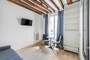 Ein Zimmer mit flachbildfernseher an der Wand, einem kleinen runden Tisch mit zwei Stühlen, einer Couch und einem Fenster mit Vorhängen.