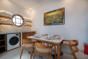 Een eetruimte met een tafel gedekt voor vier personen, omringd door houten stoelen met geweven zittingen. Achtergrond: een wasmachine, en aan de muur hangt een schilderij.