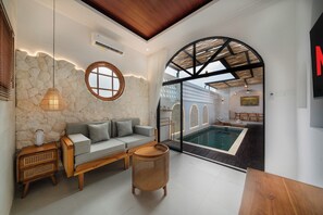 A modern interior with a stone wall, a round window, a sofa, a wicker chair, and a view of an outdoor swimming pool area. - Kanoto Villa by Betterplace (Jimbaran)