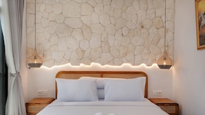 Quarto de hotel com cabeceira de padrão em pedra, duas luminárias de apoio, uma cama bem arrumada e um quadro na parede.