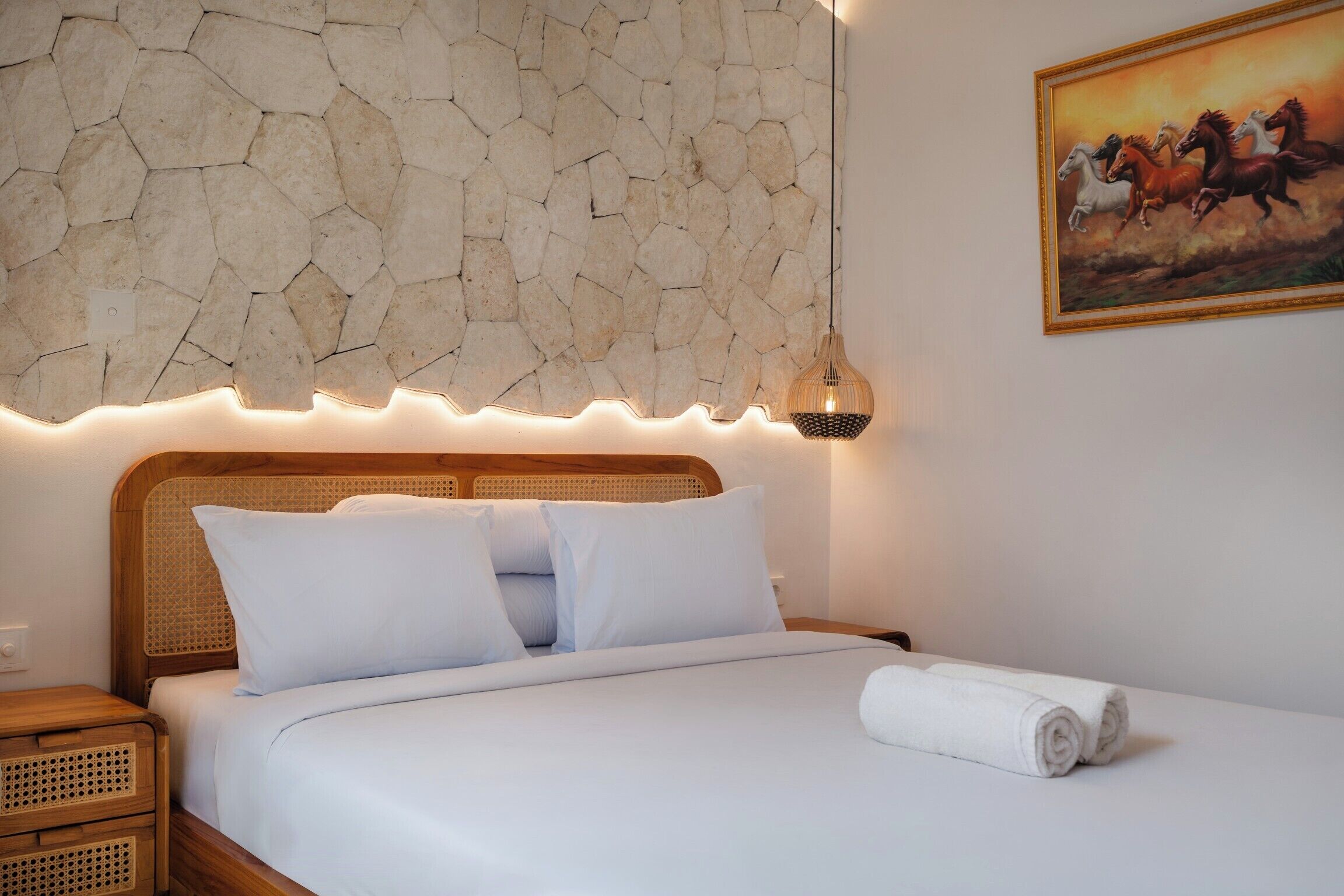 A bed with white linens, a stone wall, a painting of horses, and a bedside lamp.