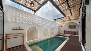 Uma área de piscina com vista desobstruída para o céu, um espaço de estar com mesa e cadeiras, e uma parede com aberturas circulares.