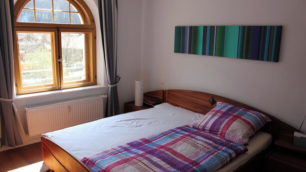 Ein Schlafzimmer mit einem Bett, einem Fenster, einem Heizkörper, einem Nachttisch und einem an der Wand befestigten Kunstwerk.