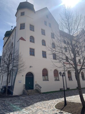 A white building with multiple windows and a green door, featuring a stone-paved courtyard and a few trees. - Vacation Rental: 65 square meters, separate bedroom and balcony, Apt. 7 (Aschau im Chiemgau)