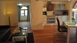 Eine moderne Küche mit Edelstahlgeräten, ein Essbereich mit Glastisch und ein Wohnzimmer mit dunkler Couch und Glastisch.